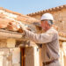 Un artisan inspecte la toiture et les murs d’une maison ancienne en rénovation thermique, dans une ambiance lumineuse.